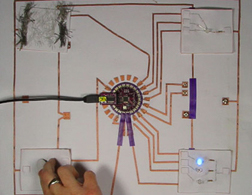 A large fabric breakout board with a Lilypad in the middle and multiple copper fabric traces linking it to various input and output swatches