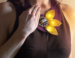 A person wears a DIY felt flower necklace embellished with a circular microcontroller. A hand covers part of the board