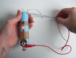 The left hand holds a DIY knitting nancy loom with an LED and copper tape while the right hand pulls a knitted sensor to activate the LED