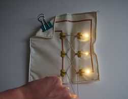 A hand pulls the yarn emerging at the bottom of a 3x3 inch fabric swatch with three LEDs illuminated