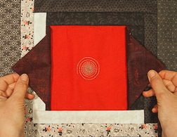 Two hands opening two triangle sides to a red quilt patch with a center silver spiral