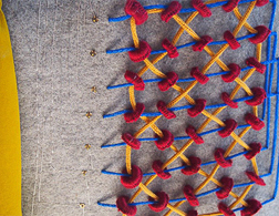 A matrix of red rings adorn a gray sheet of felt. Blue and yellow knitted cords are woven through the rings