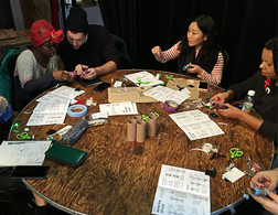 4 people around a table helping each other build a knitting loom